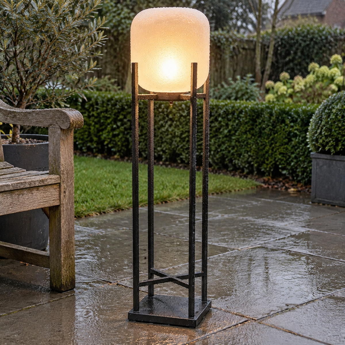 Standing LED metal lantern floor lamp lit on wet paving in a British back garden