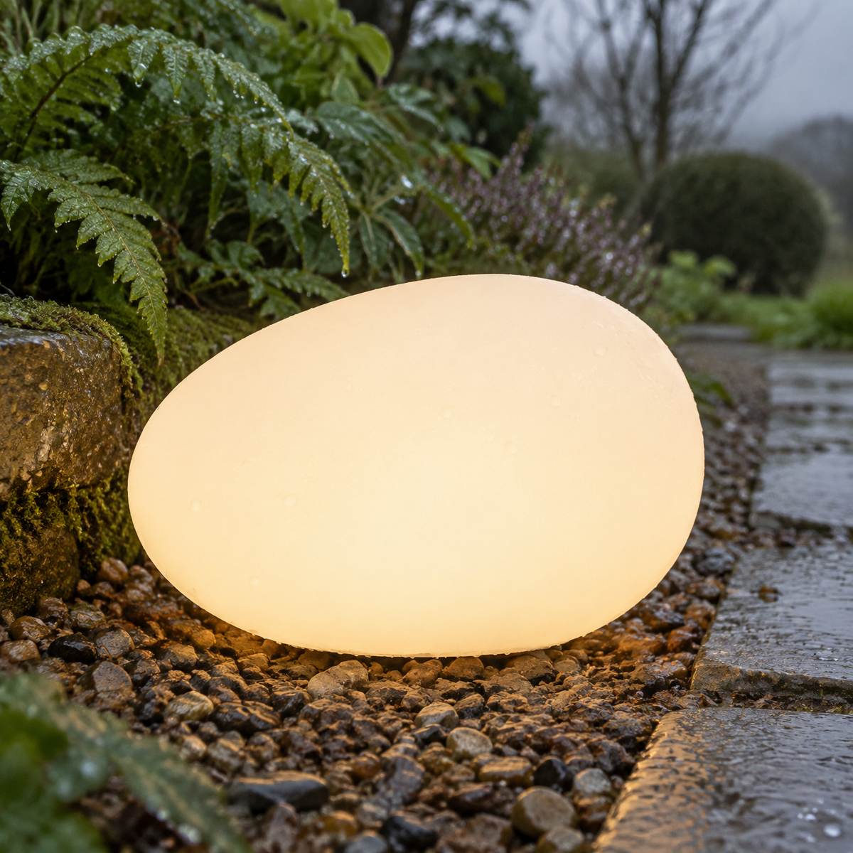 Pebble-shaped waterproof garden lamp glowing on wet gravel beside a path