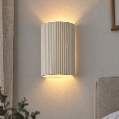 Scandinavian beige plaster oval wall sconce lit above a bed on a chalky painted wall