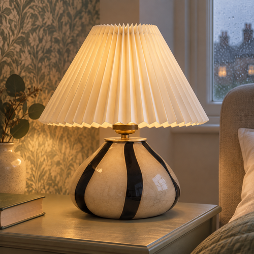 Pleated ceramic bedside table lamp switched on beside a hardback book