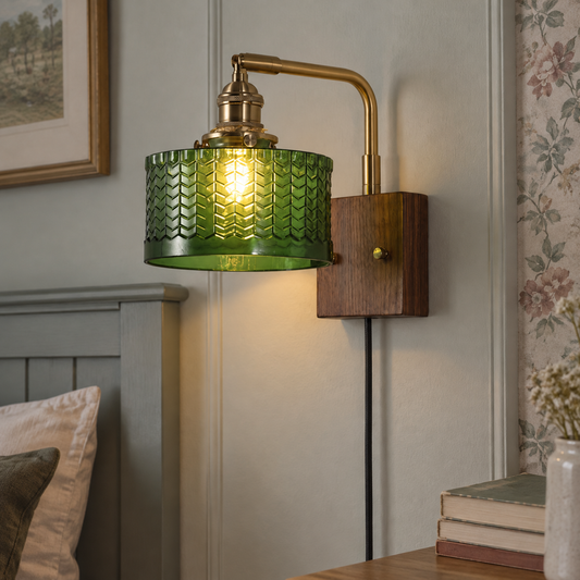 Brass and ribbed glass plug-in wall sconce mounted beside a bed, its arm projecting from the wall and flex running down to a socket, lit warm in a British bedroom