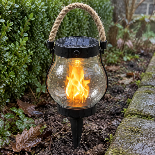Solar flame torch light staked upright in a garden border beside a box hedge, its amber flame effect flickering on a drizzly grey afternoon