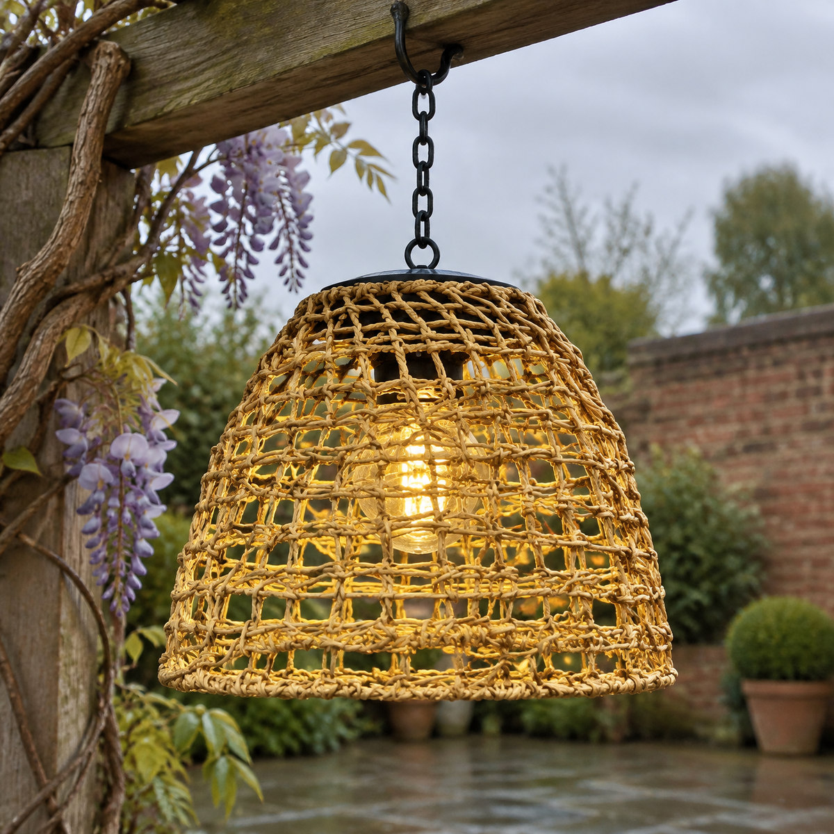 Bell-shaped solar hanging lantern suspended from a weathered pergola beam over a patio, glowing warm white at dusk