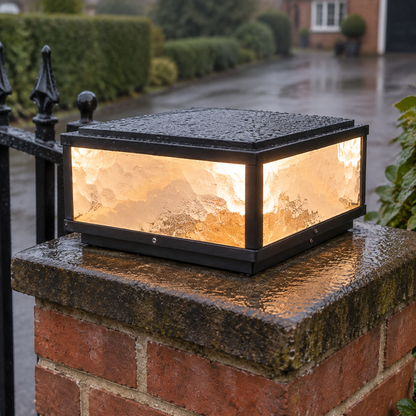 Large square die-cast post cap pillar light standing on a rain-darkened brick gate pillar beside a wrought iron gate