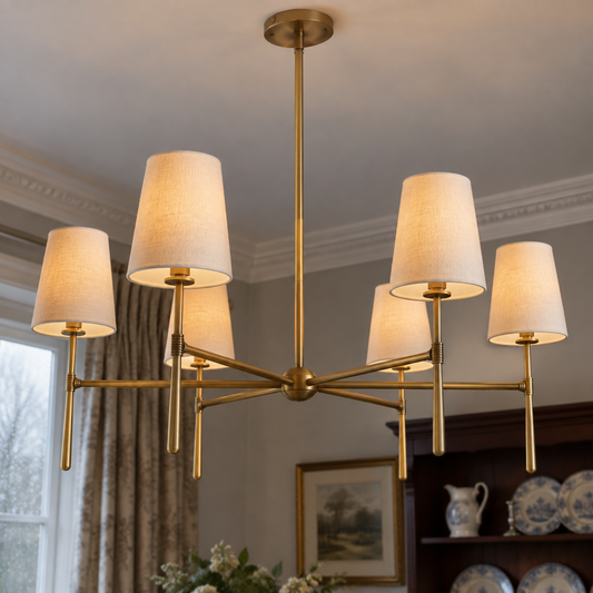 Round brass chandelier ring hung with cylindrical linen shades above a dining table, shades facing down and lit warm