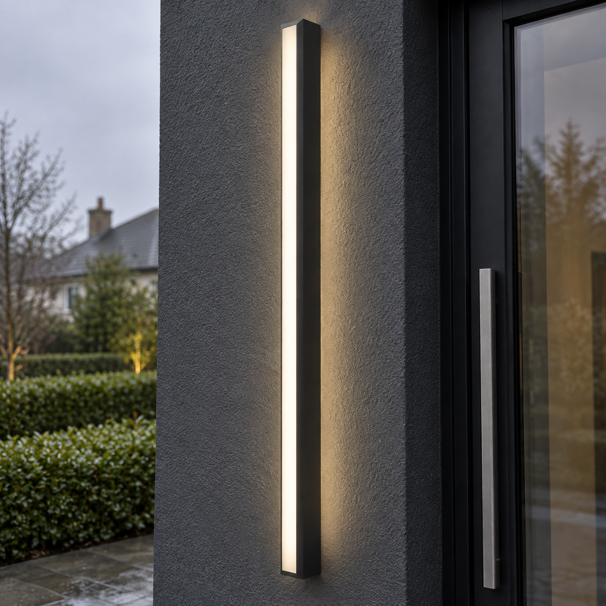 Long slim black linear LED wall light mounted upright on charcoal render beside a glazed front door at dusk