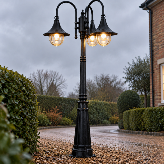 Vintage black outdoor lamp post standing upright on a wet gravel driveway, flared lantern head lit warmly beside clipped hedging