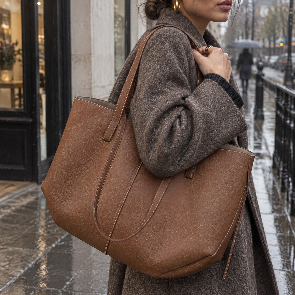 Large leather everyday tote bag carried over a woman's shoulder on a drizzly pavement outside a shop doorway